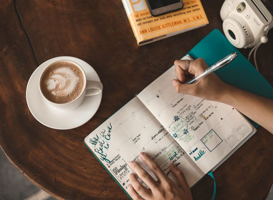 A person writing a plan in a notebook next to a coffee